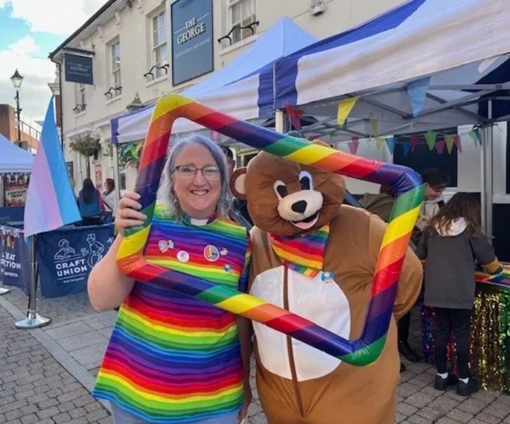 Woman in rainbow top posing with the Healing Hubs bear mascot at a Pride event