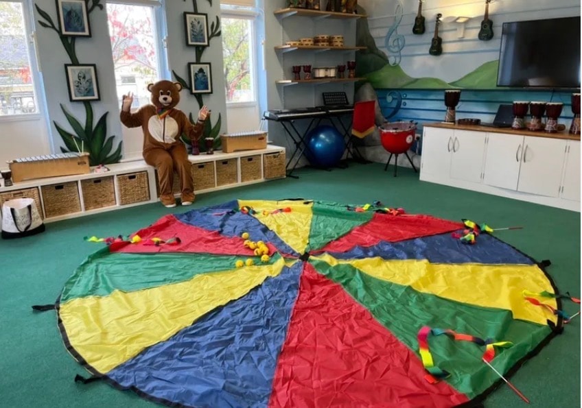 Healing Hubs bear mascot in a bright activity room with a colourful parachute laid out on the floor