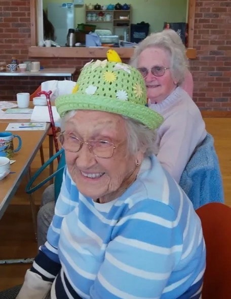 Two elderly women smiling at a community gathering, one wearing a decorated green hat