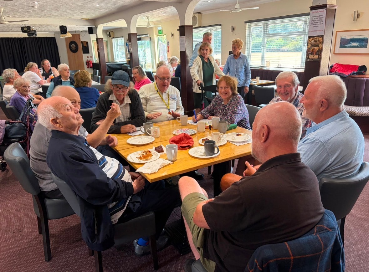 Busy community room full of members chatting over tea and cake at a Healing Hubs event